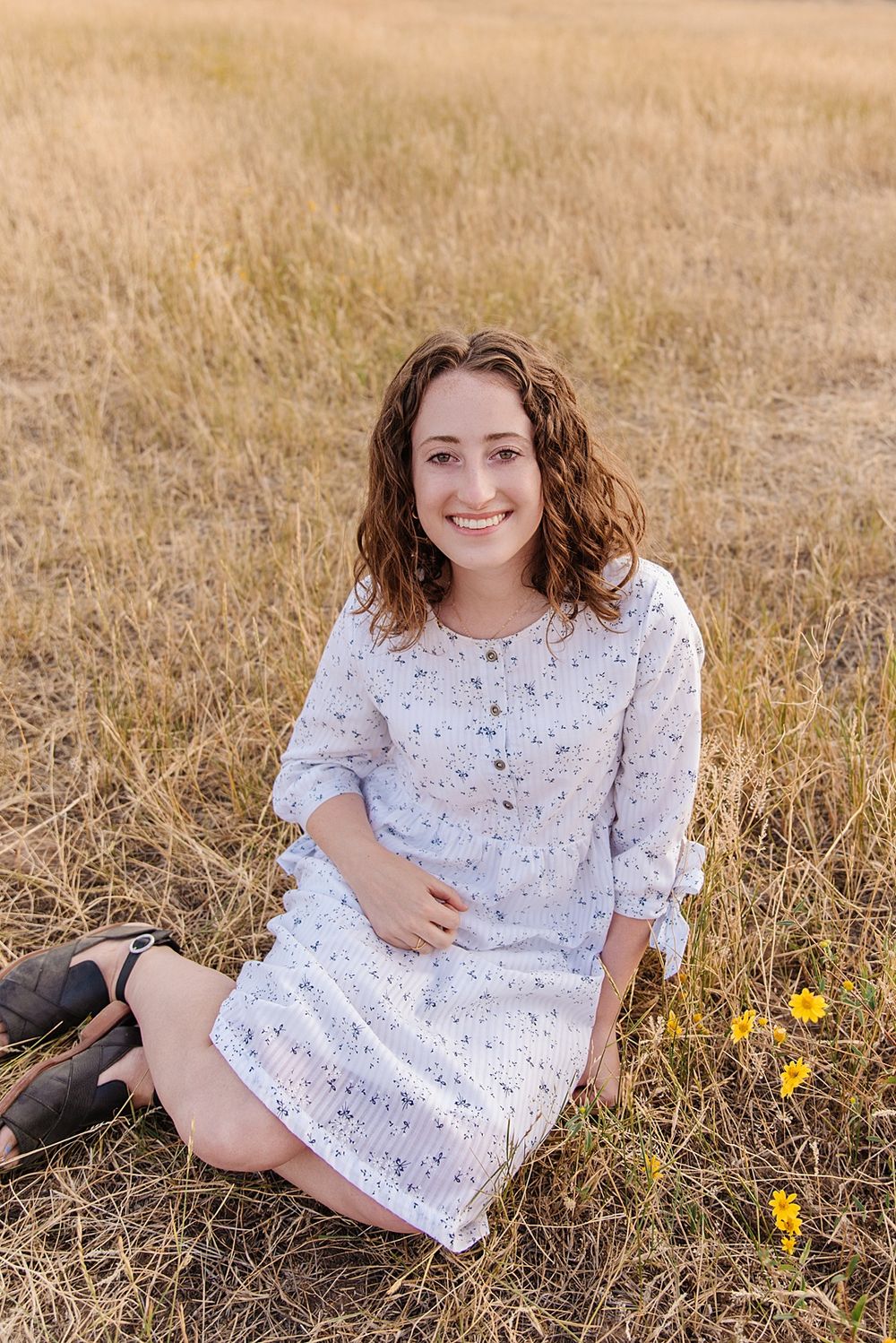 Person in white dress sitting in golden grass field with small yellow wildflowers.
