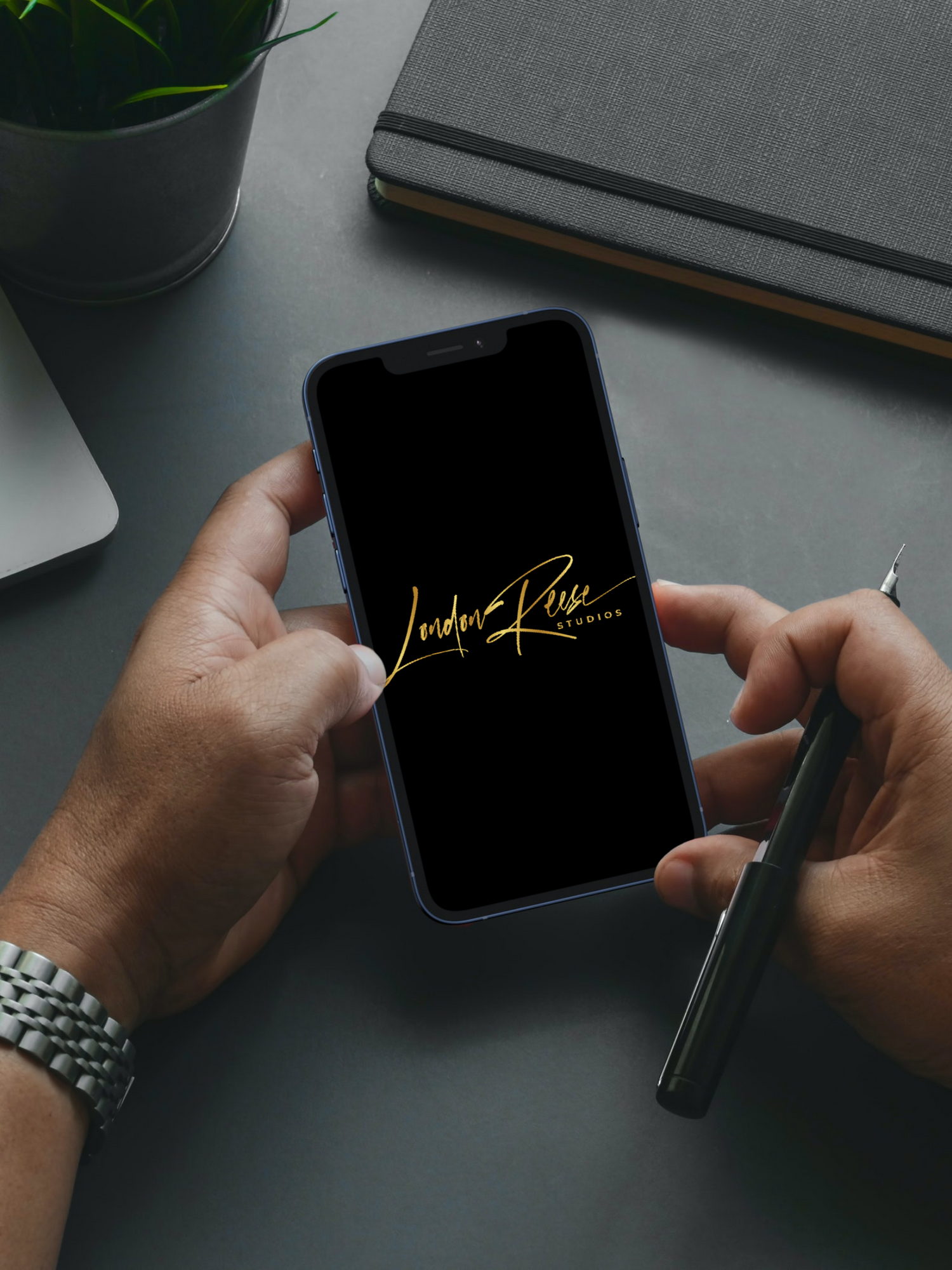 Hands holding smartphone displaying gold signature logo on dark background with notebook and plant visible.