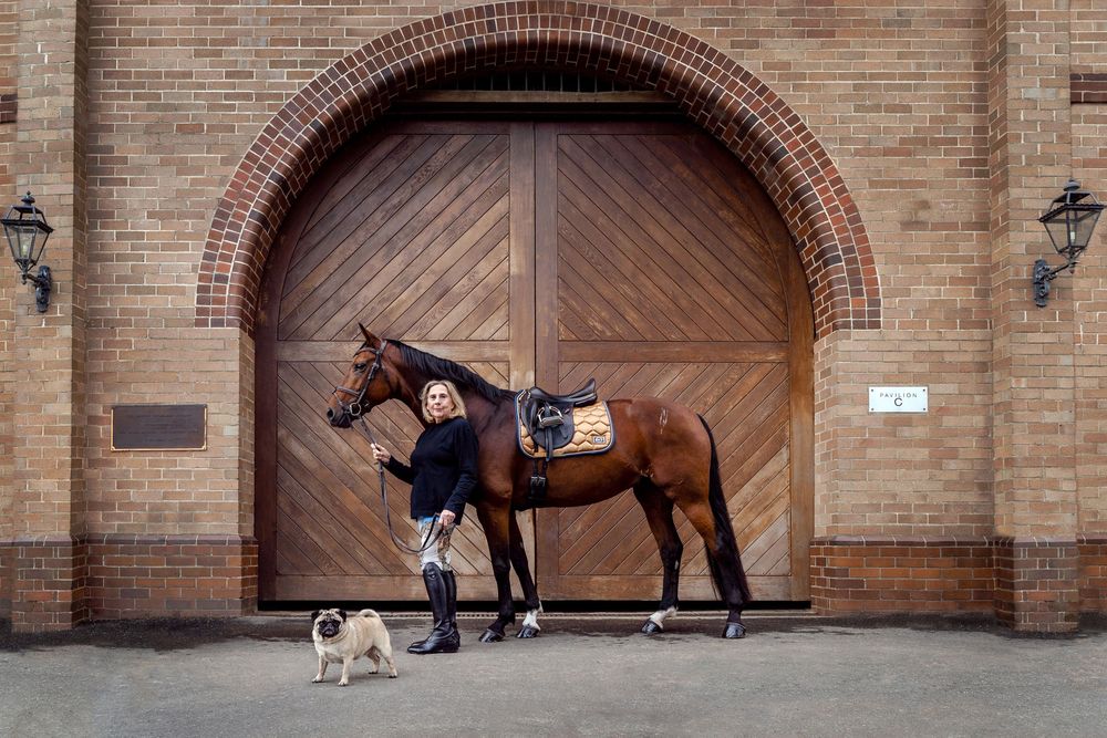 Woman stands with a brown horse and pug dog in front of large wooden doors in Centennial Parklands Equestrian Centre photoshoot