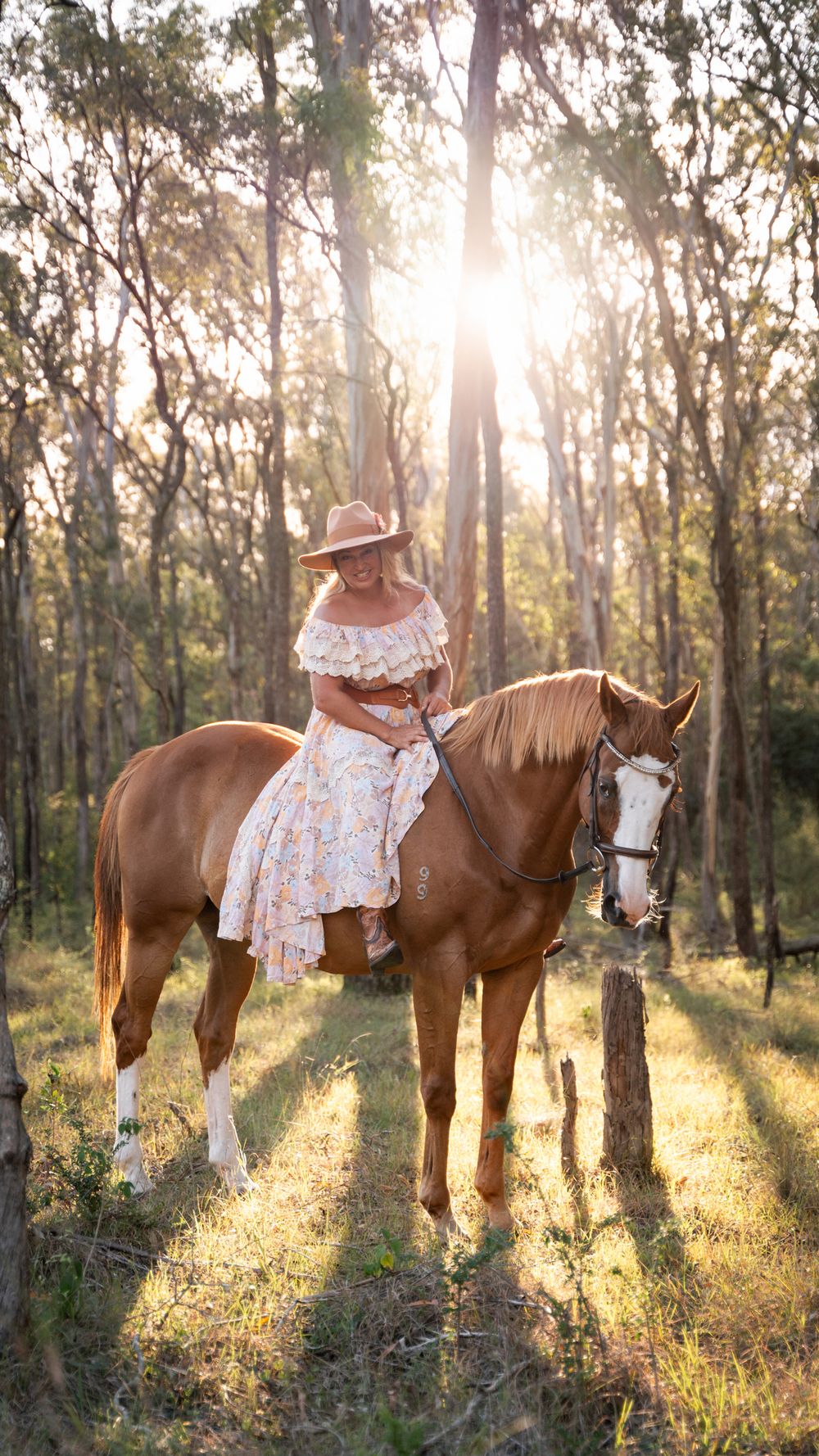 Woman in floral dress and cowboy hat riding a chestnut horse in a sunlit forest clearing in Scheyville national park Maraylya