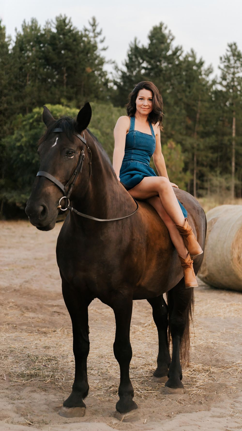 Woman in blue dress and brown boots sitting sidesaddle on a dark brown horse in a rural farm setting in sydney