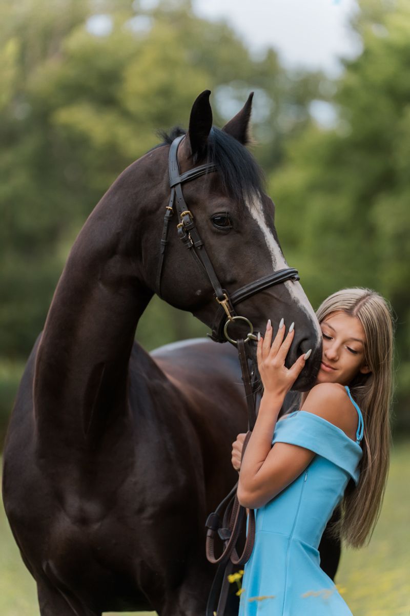 A young blonde woman in a blue off-shoulder dress tenderly embraces a dark bay horse outdoors in Sydney Terry hills