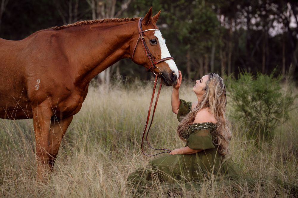 woman in a dress kneels in tall grass, tenderly facing a chestnut horse with a white blaze in a natural setting in Scheyville national park