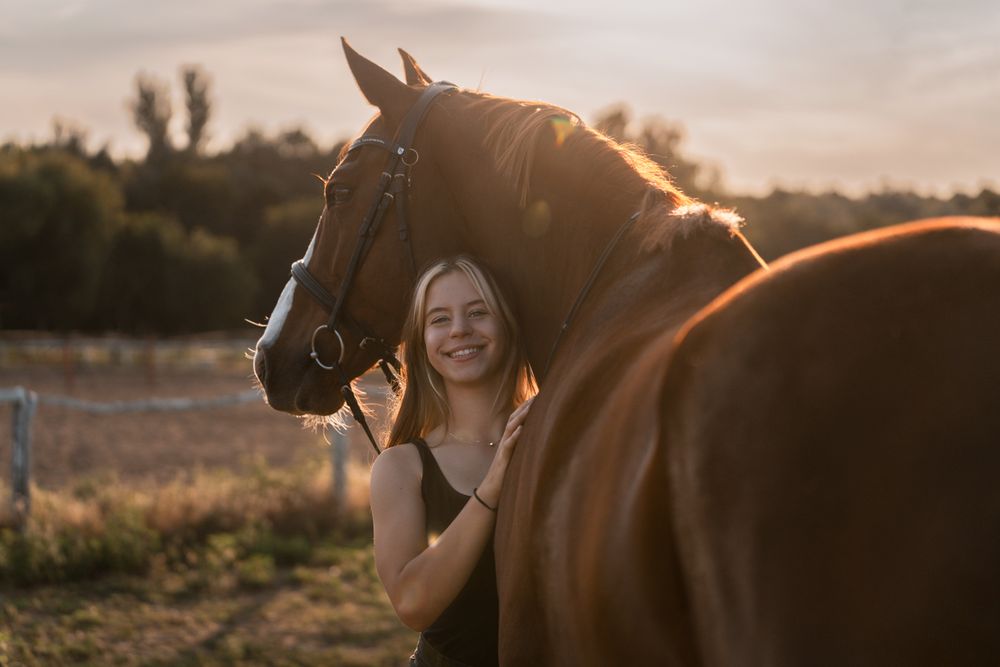 Young blonde woman smiling beside a chestnut horse in golden sunset light on a rural farm in Hunter Valley horse photoshoot