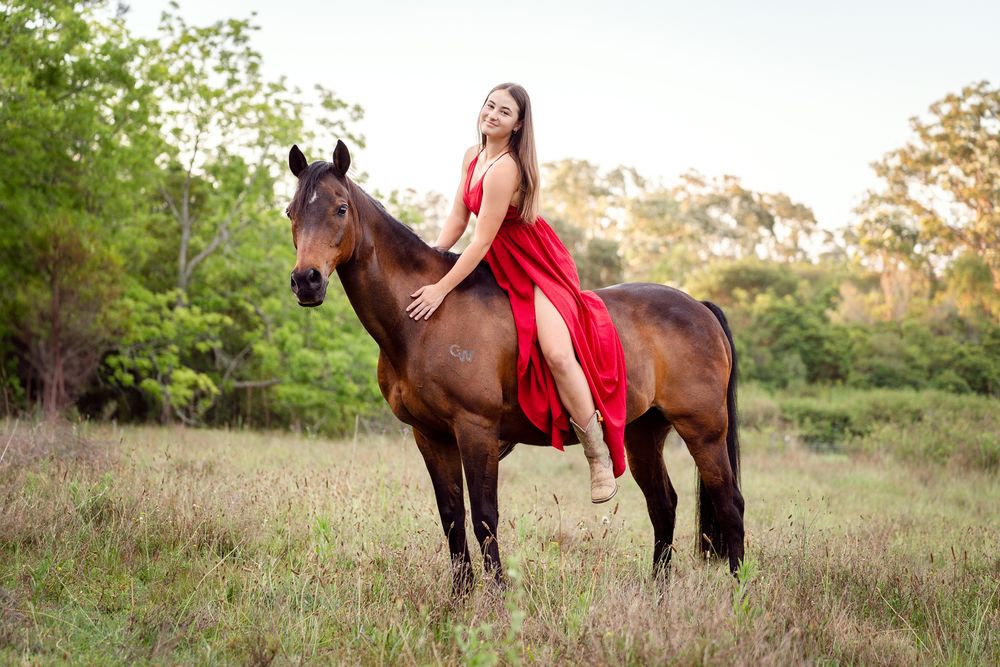 Woman in red dress riding a brown horse in a green field with trees in the background arcadia photoshoot with a horse