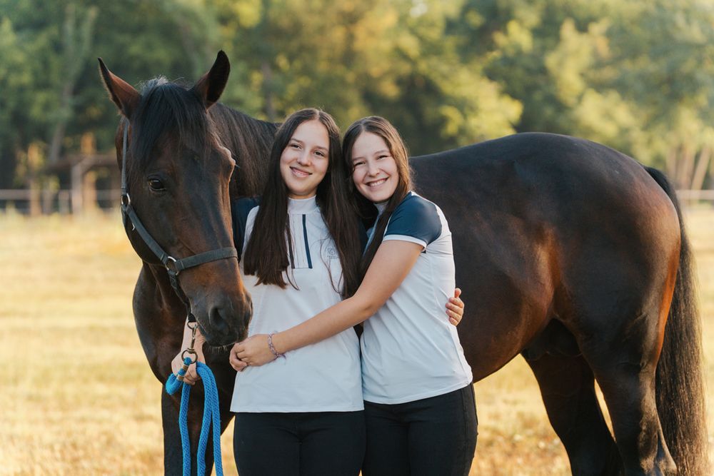 sisters pose with a dark brown horse in a sunny field, one holding a blue lead rope. during family photoshoot with a horse in Camden sydney