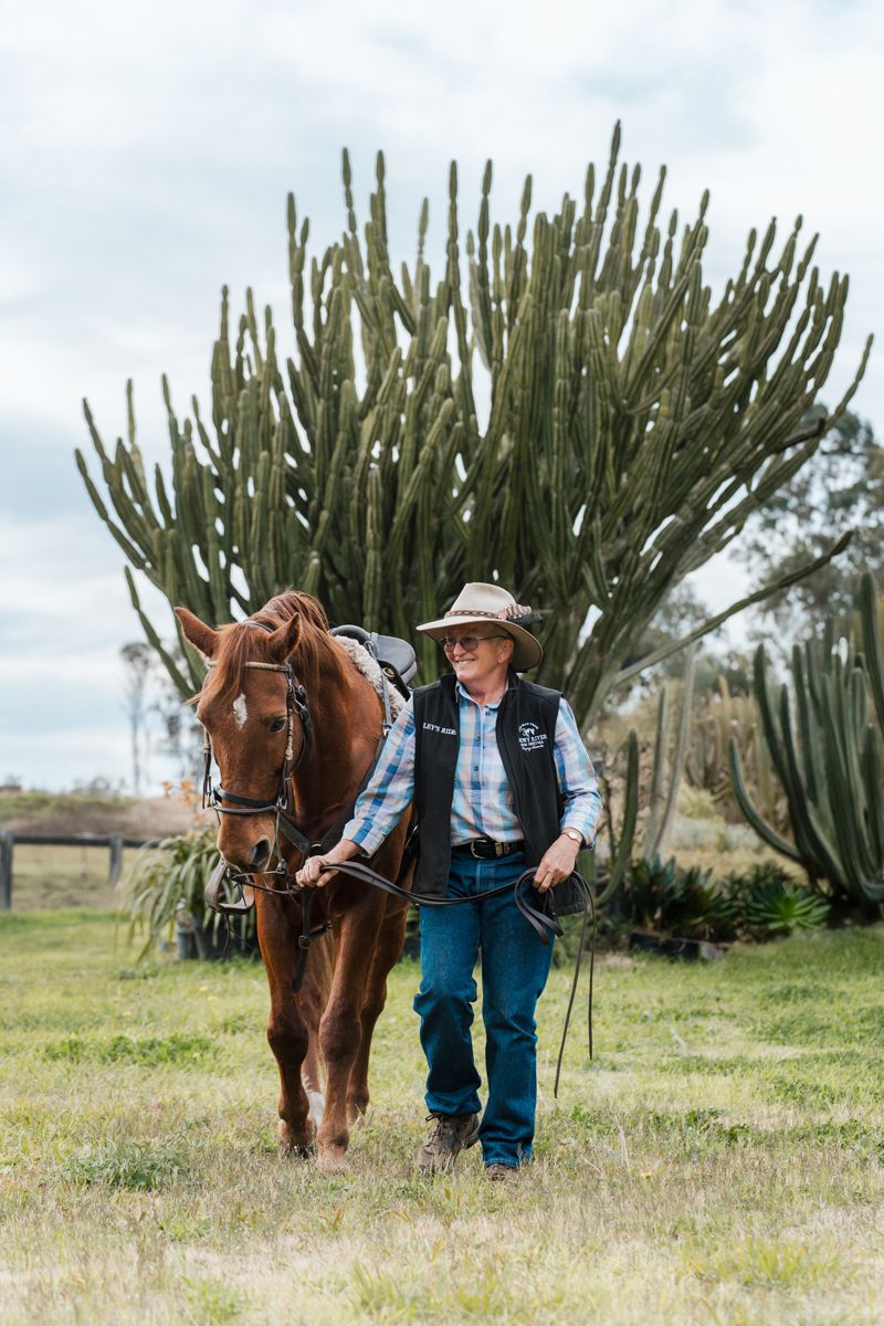 A cowboy in a hat and vest walks alongside a chestnut horse near a tall cactus in a green field portrait photoshoot with a horse in Cattai