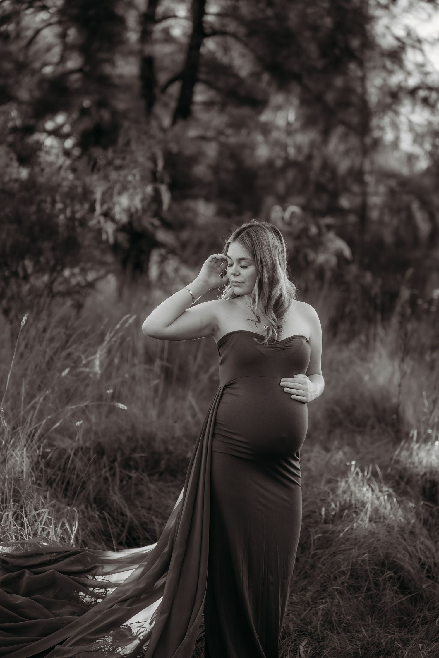 Pregnant woman in elegant strapless gown poses outdoors in black and white maternity portrait among tall grass.