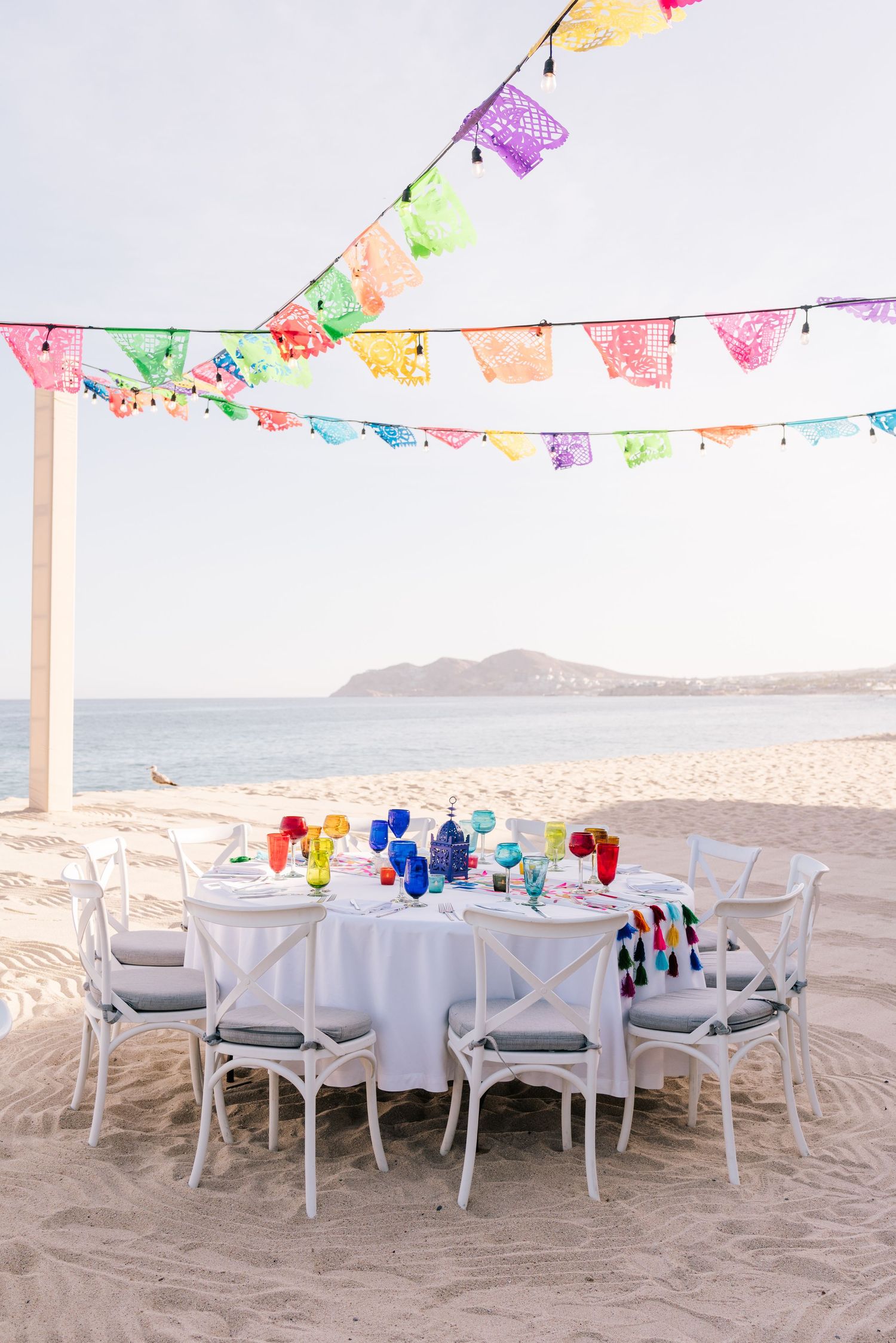 Colorful papel picado banners over a white table set on a sandy beach with ocean views for a festive celebration.