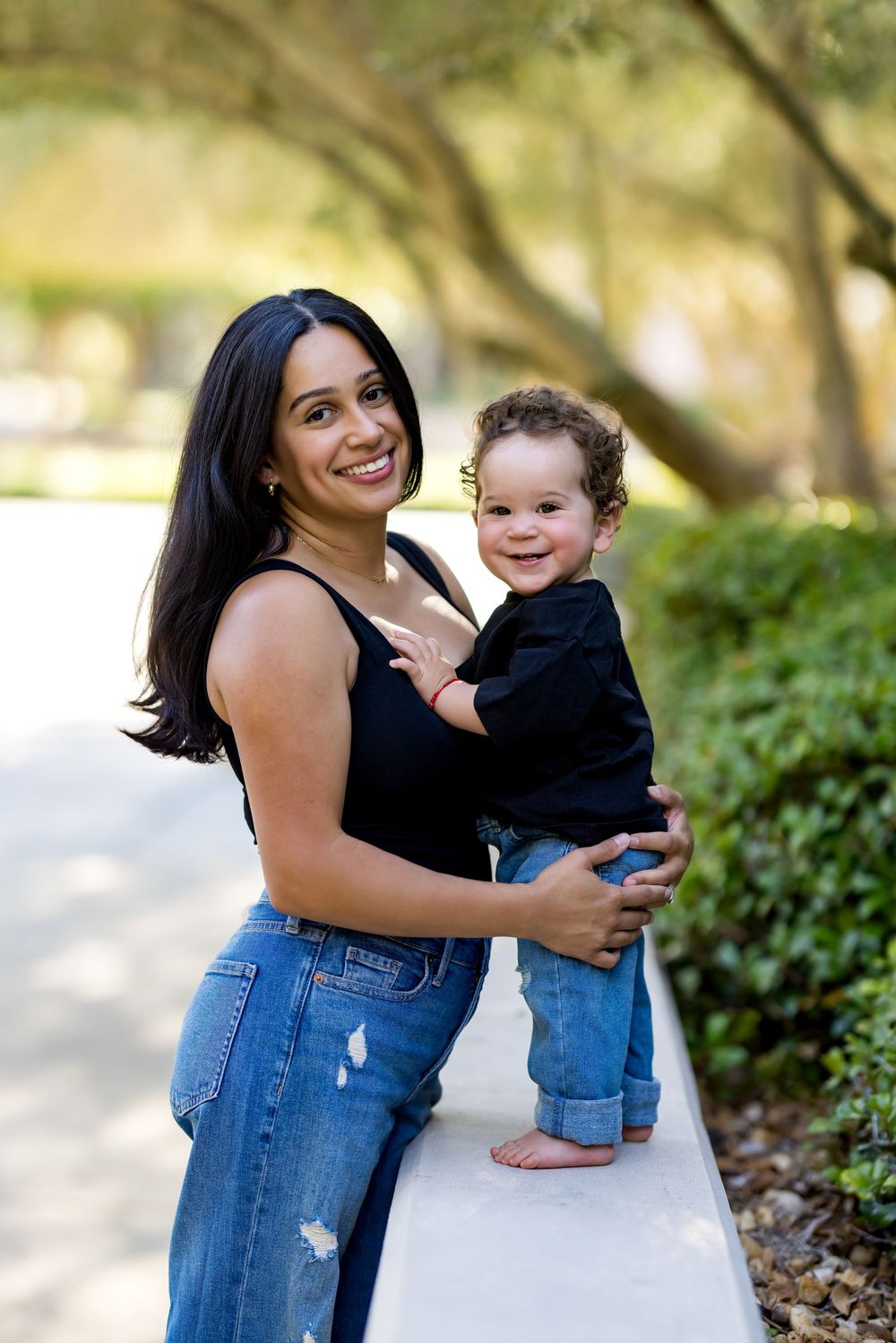 Smiling mother holding toddler boy in black shirts and jeans on white wall outdoors in park setting.