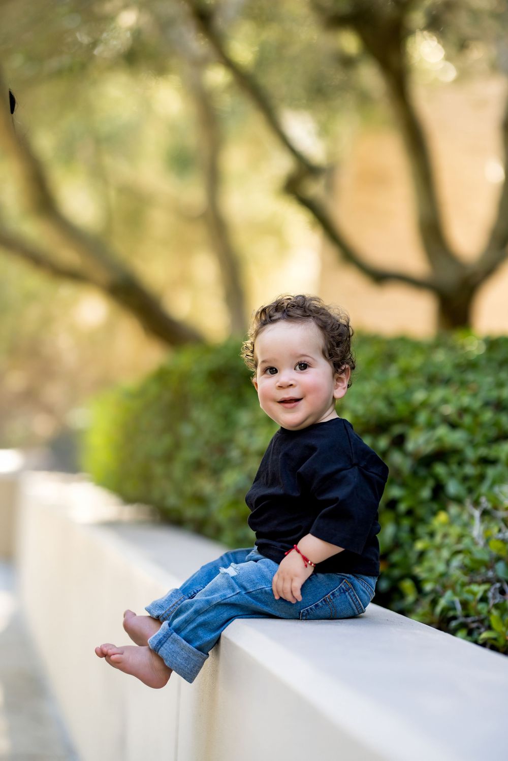 Cute toddler boy in black shirt and jeans sitting on white wall in sunny outdoor park setting.