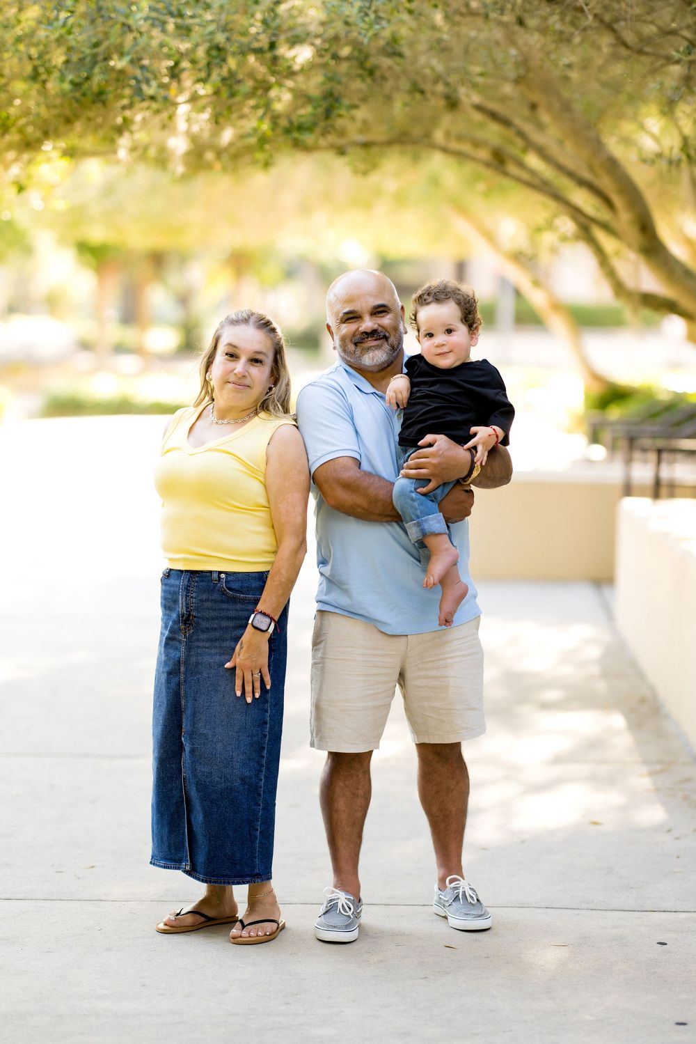 A smiling family of three poses outdoors, with a man holding a baby and a woman in a yellow top and denim skirt.