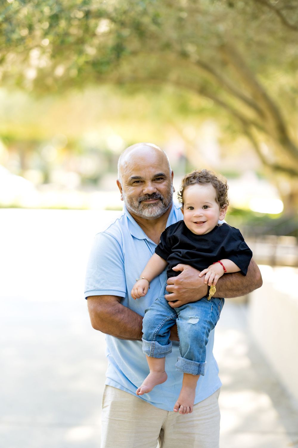 A bald man with a beard holds a smiling toddler in a sunny park, both wearing casual clothes.