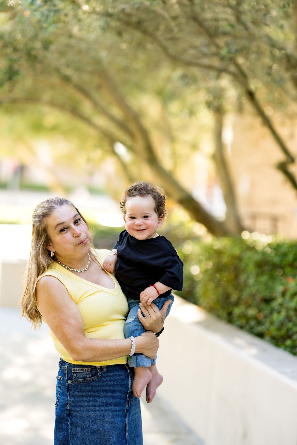 Woman in yellow top holds smiling baby in black shirt outdoors near trees on a sunny day.