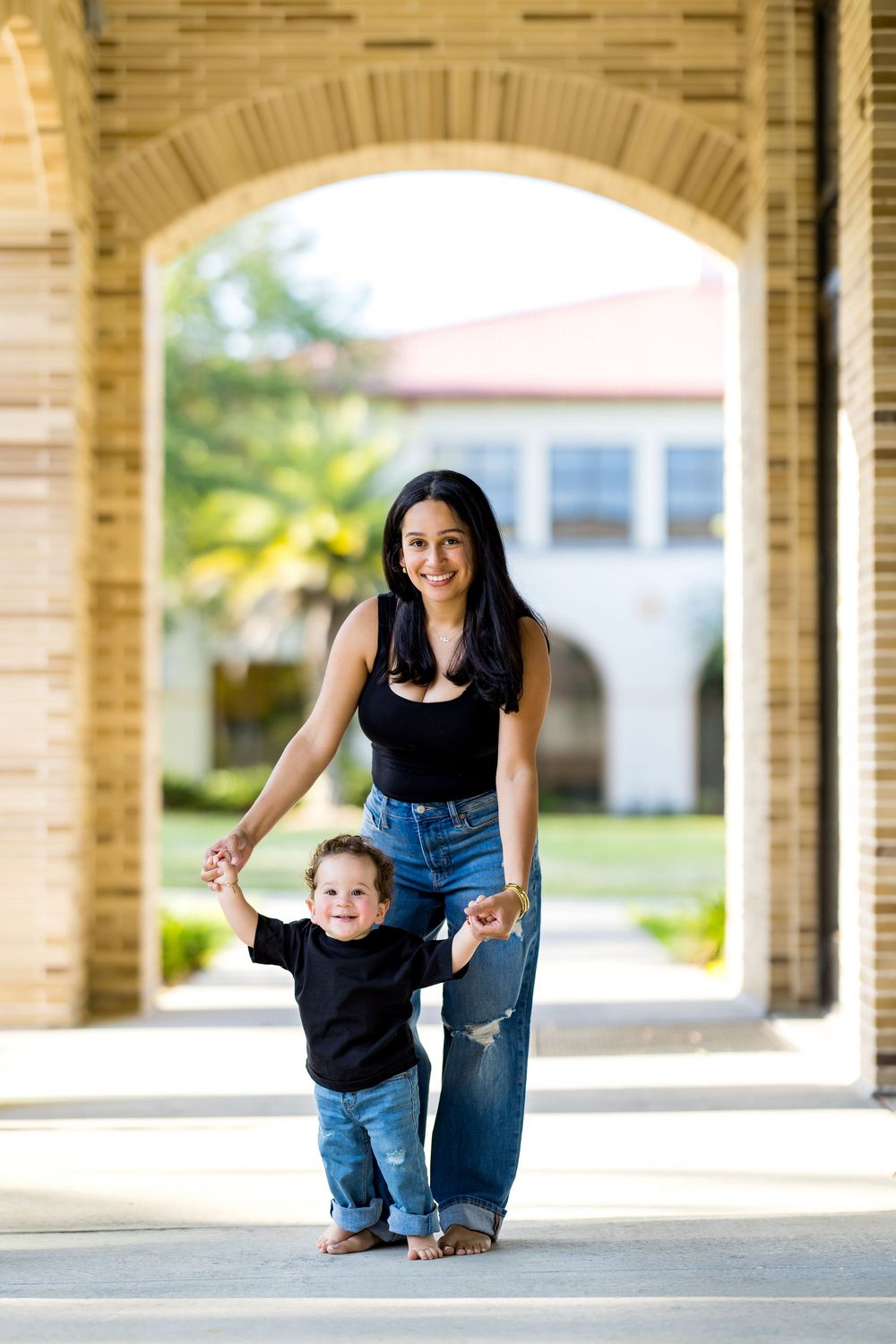 A smiling mother helps her baby take first steps under a brick archway outdoors.