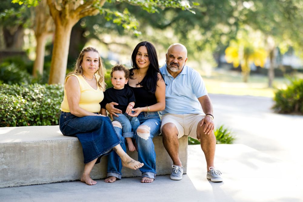 Family of four smiling together on a stone bench in a sunny park setting.
