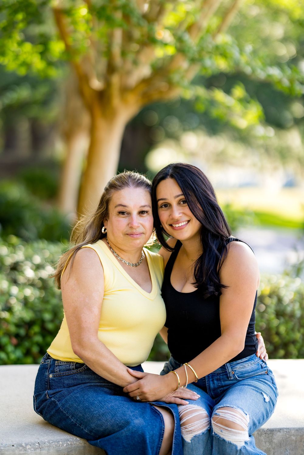 Two women smiling and sitting together outdoors on a stone bench surrounded by lush green trees and shrubs.