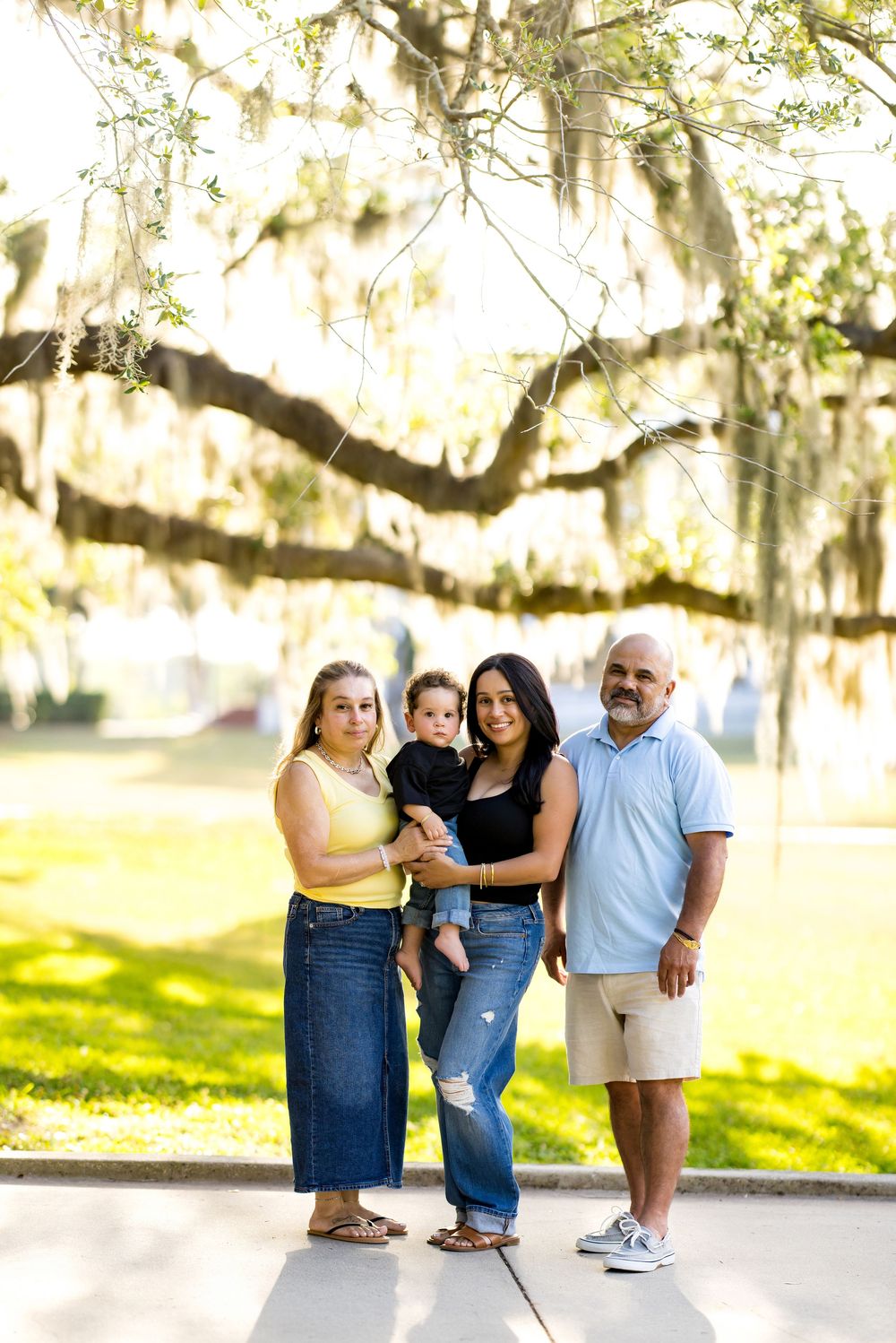Family of four posing outdoors under a large oak tree with Spanish moss in a sunny park setting.
