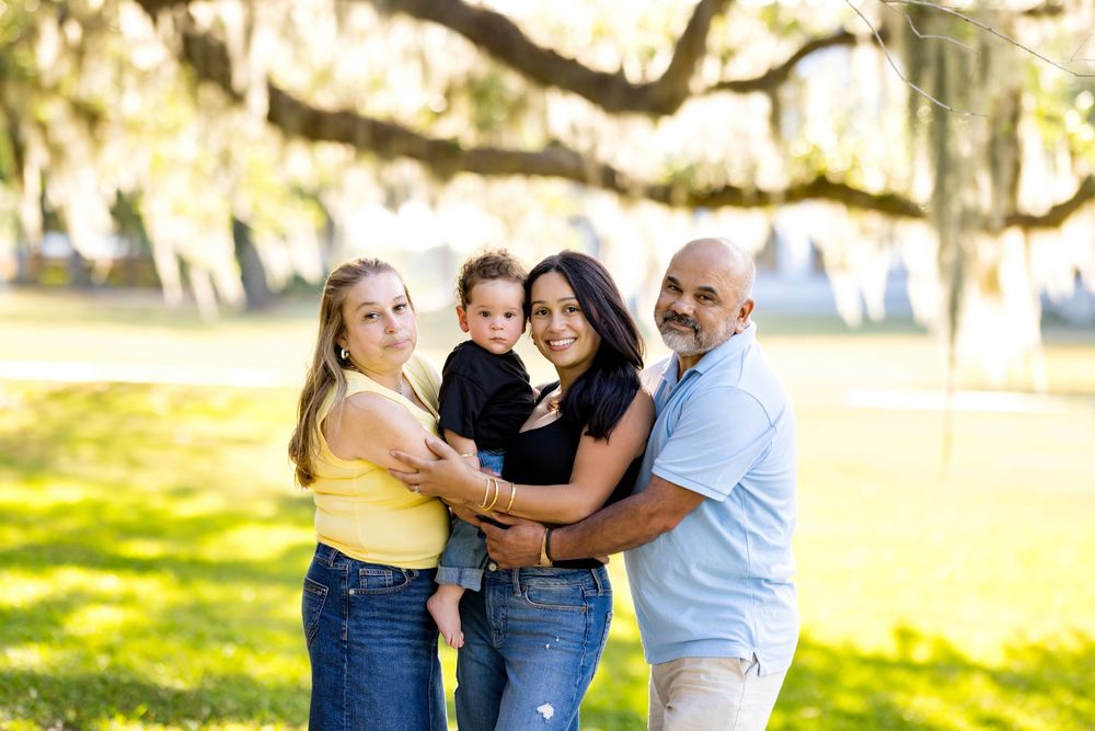 Family of four poses outdoors under moss-draped trees, parents holding toddler in a sunny green park setting.
