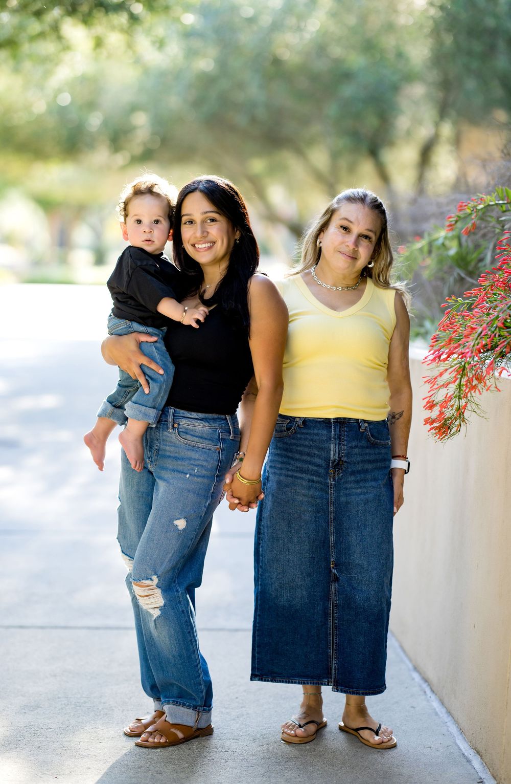 Two women and a toddler posing outdoors on a sidewalk, smiling together near red flowers.