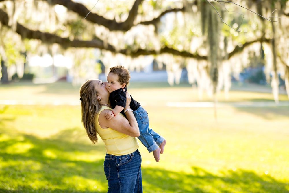 A mother lifts and kisses her young child outdoors under moss-draped oak trees in a sunny green park.