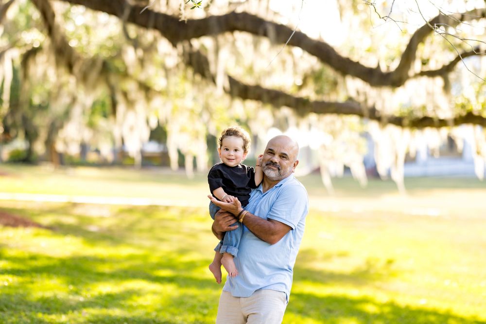 A smiling bald man holds a baby in a sunny park with sprawling oak trees draped in Spanish moss.