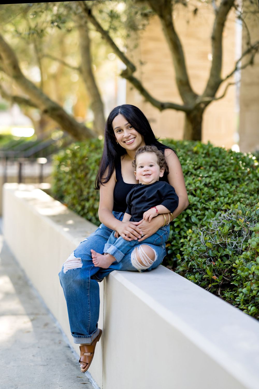 A smiling woman with dark hair sits on a wall holding a toddler, both wearing jeans and black tops outdoors.