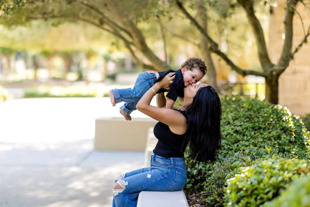 A mother sits on a bench outdoors, joyfully lifting her toddler in the air surrounded by lush green trees.
