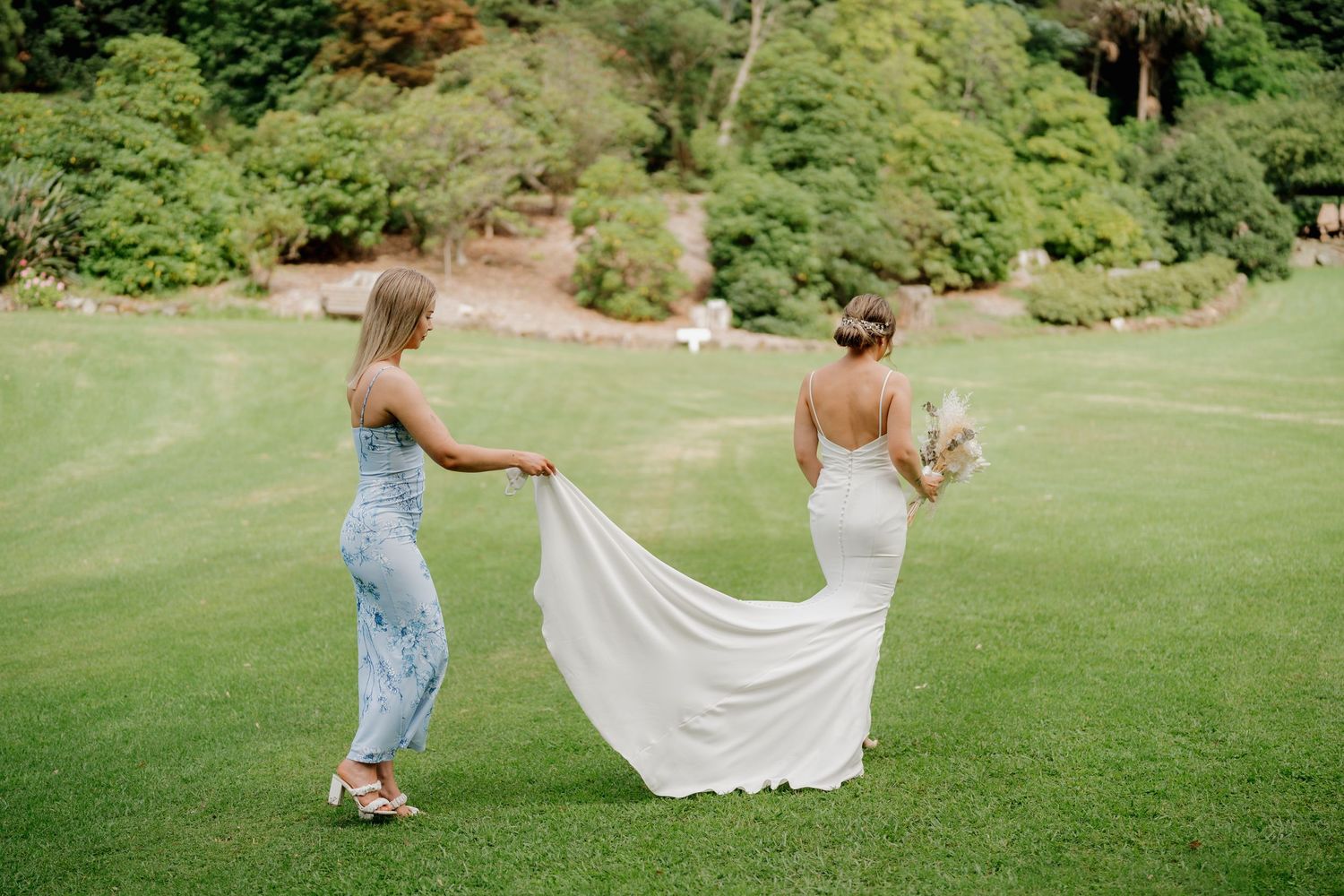 Bridesmaid in jeans helps bride with white wedding dress train on lush green lawn surrounded by trees.