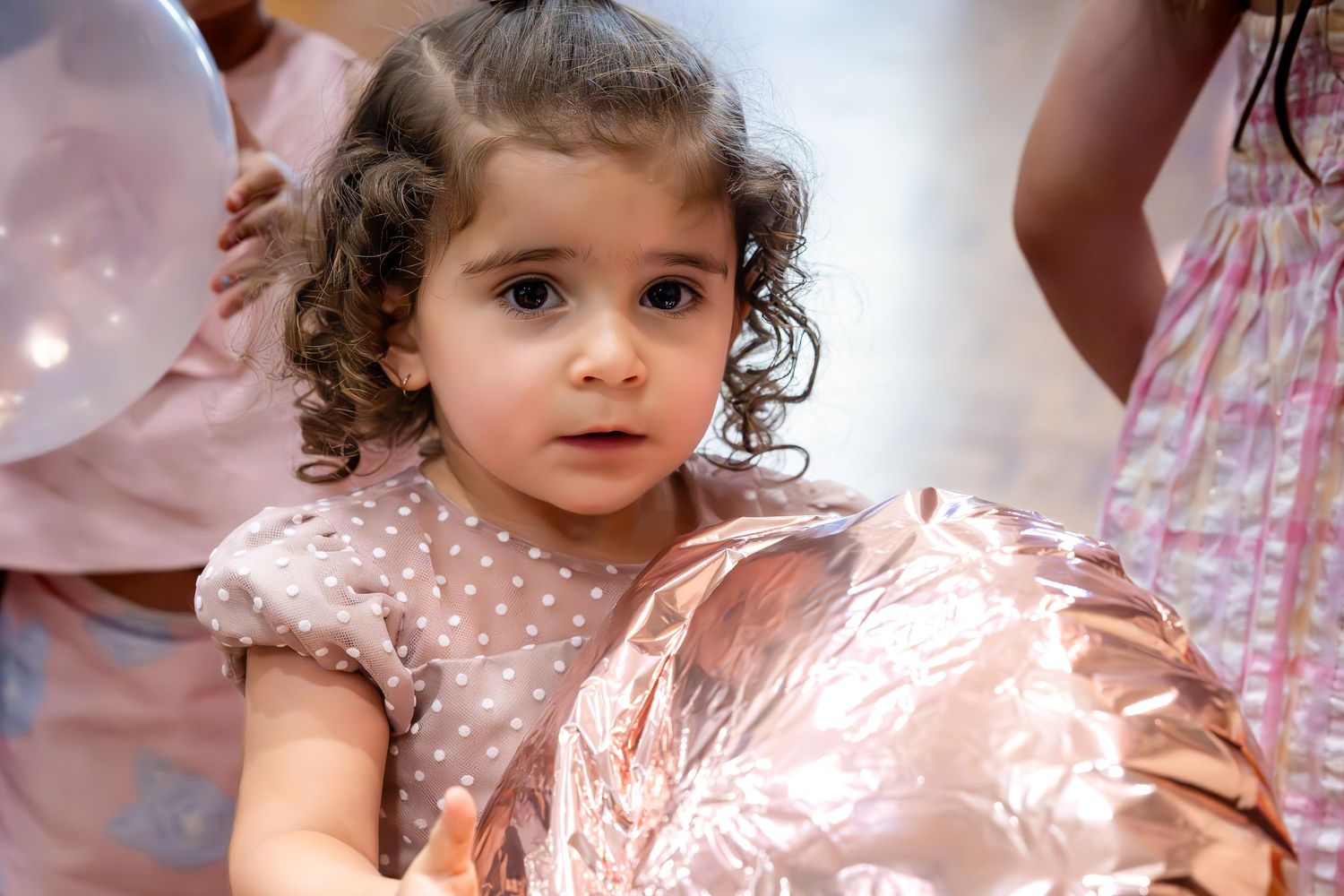 Adorable curly-haired toddler girl in a pink polka dot dress holding a shiny balloon at a party.