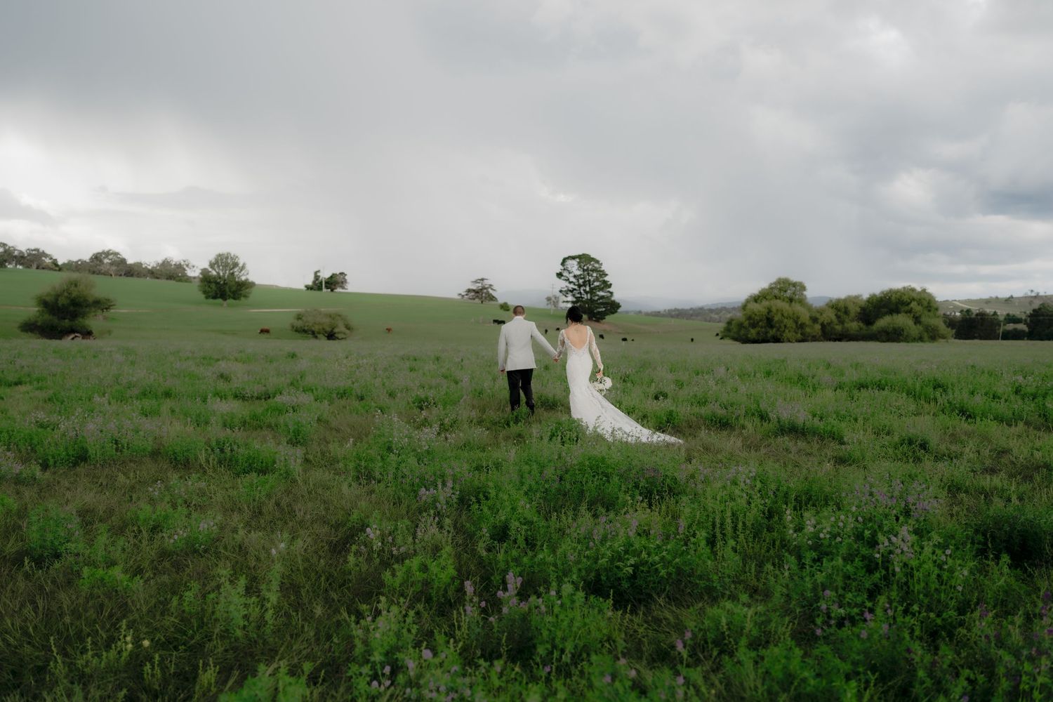 Bride and groom walking hand in hand through a lush green field under dramatic cloudy skies.