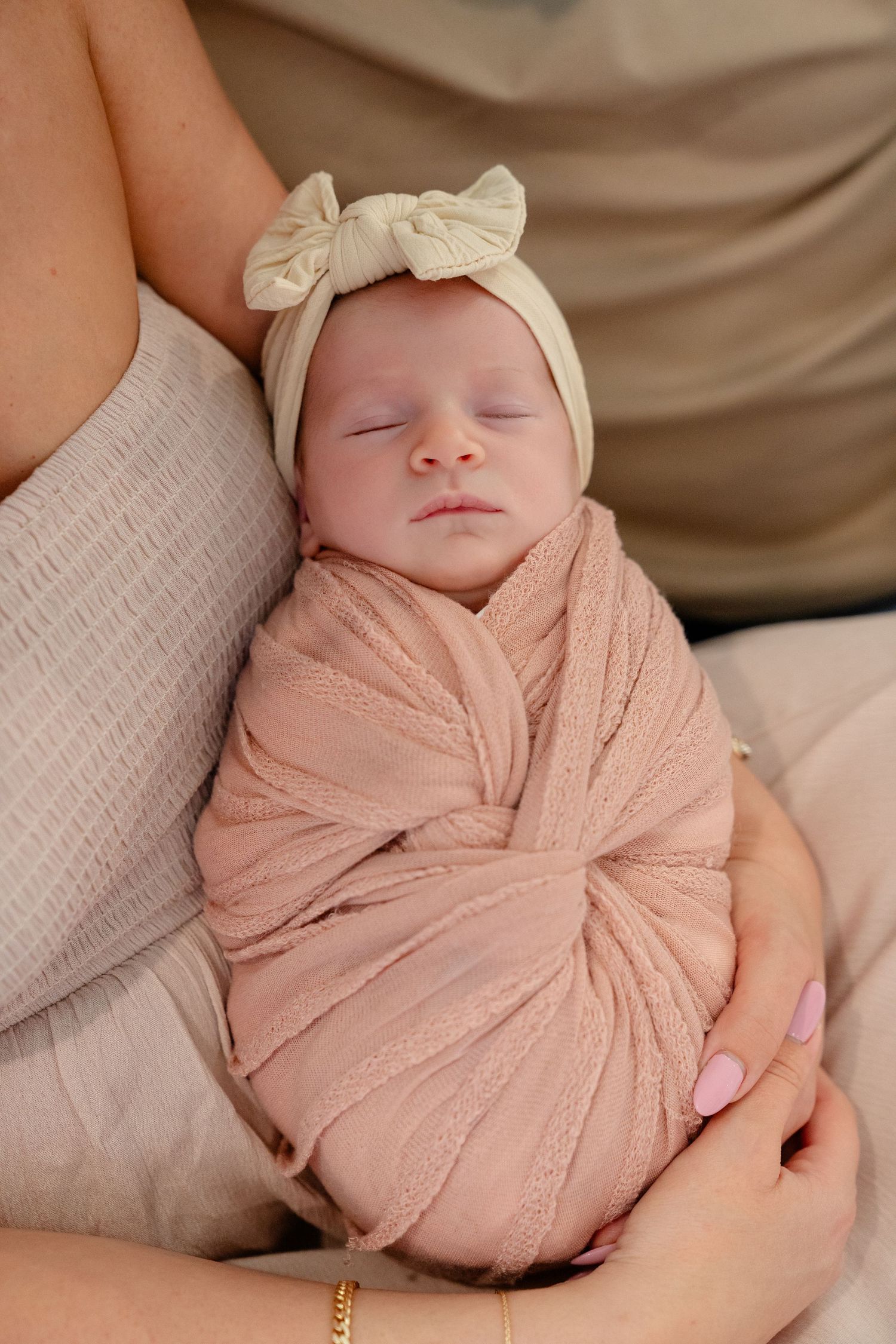 Sleeping newborn baby swaddled in pink wrap with cream bow headband, cradled in mother's arms.