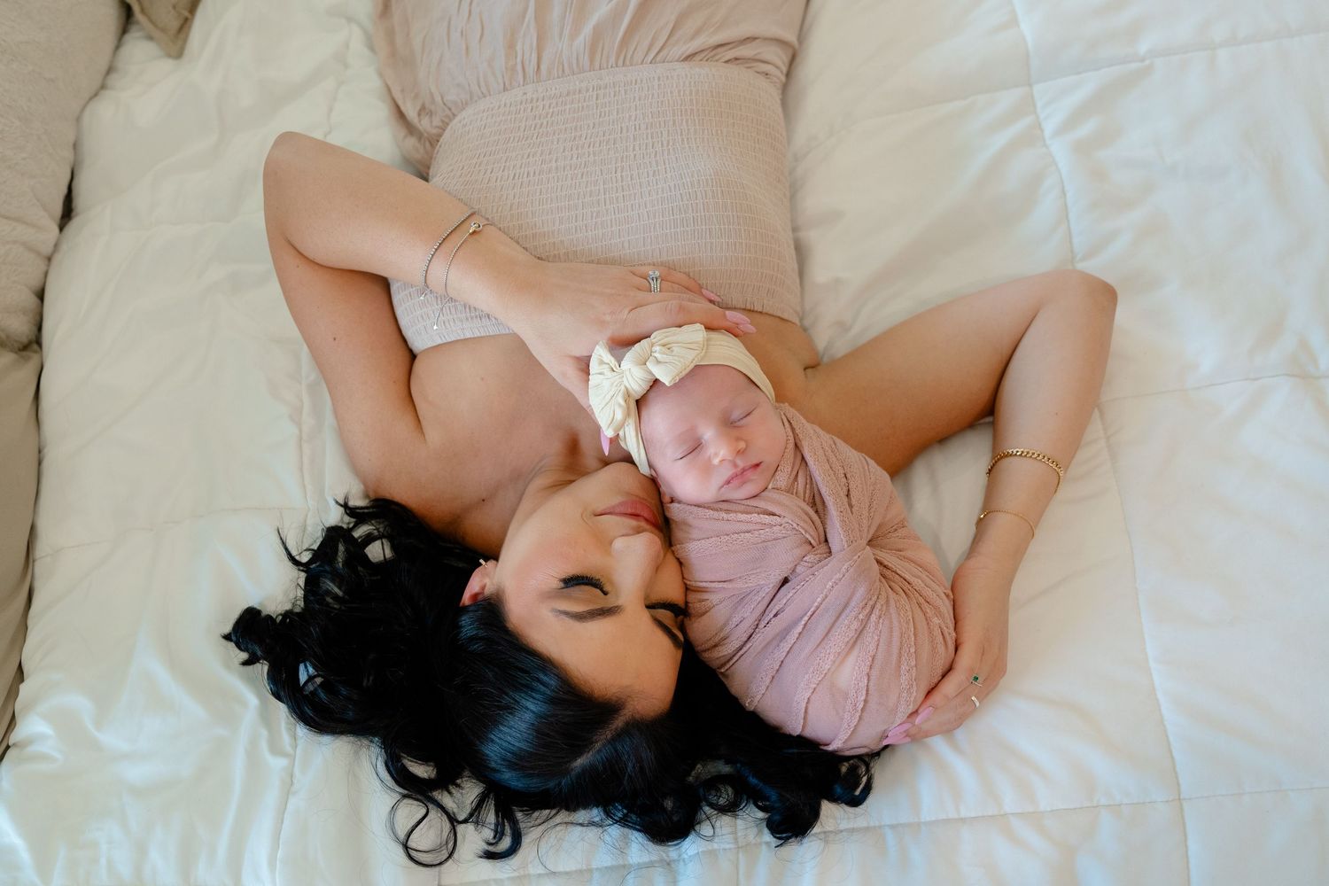 Mother lying on white bed gazing lovingly at swaddled newborn baby girl in pink wrap with floral headband.