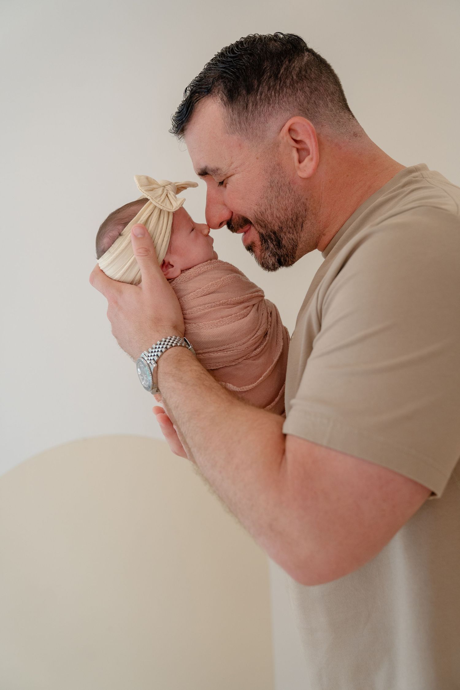 Father smiling tenderly at swaddled newborn baby held gently in his arms against neutral background.