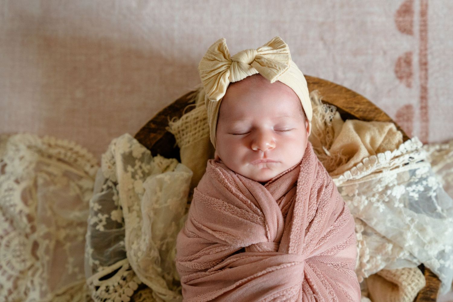 Sleeping newborn baby girl swaddled in pink wrap with gold bow headband, resting in rustic wooden bowl.