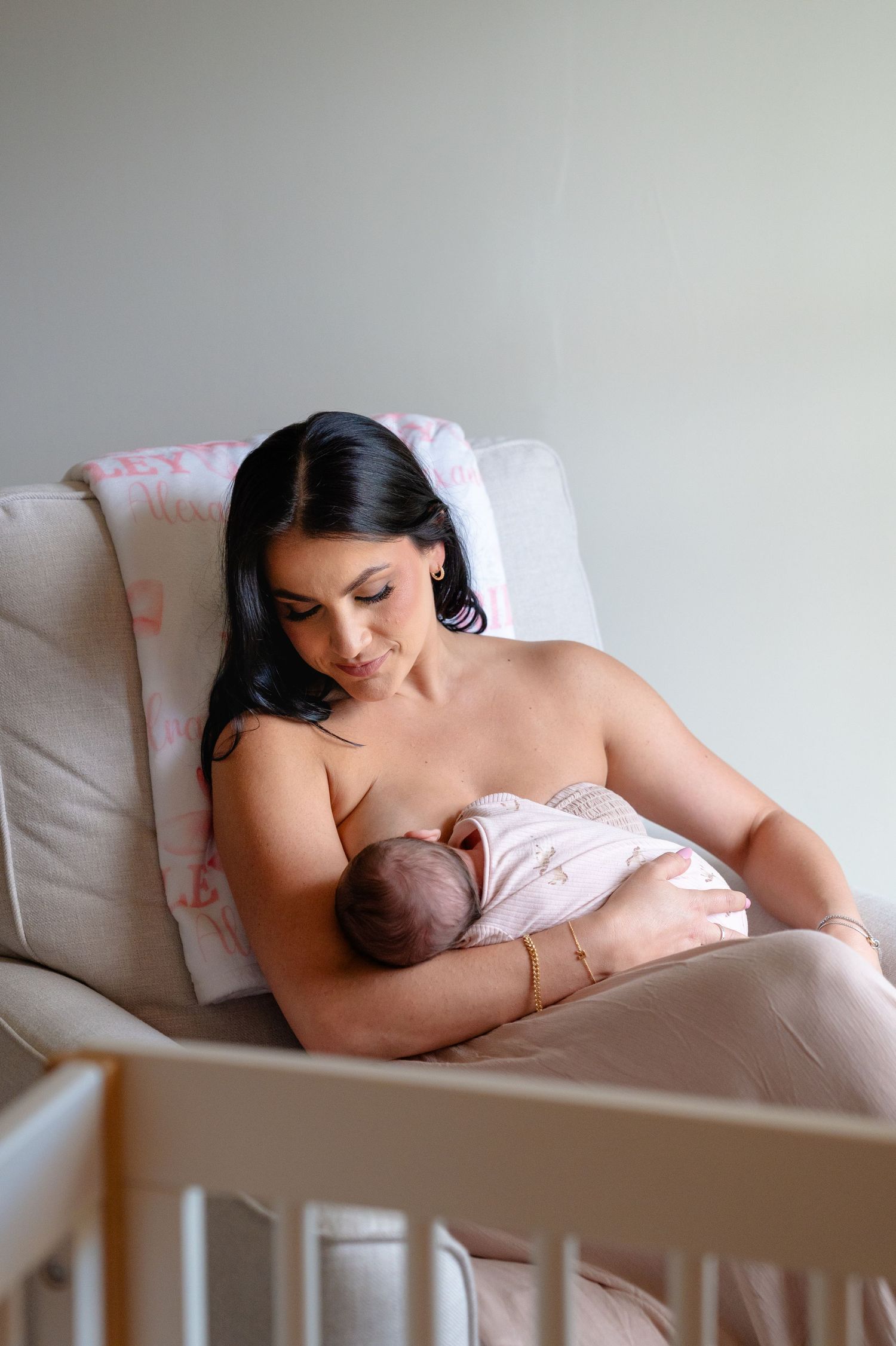 A mother breastfeeding her newborn baby while seated in a cozy armchair next to a white crib.