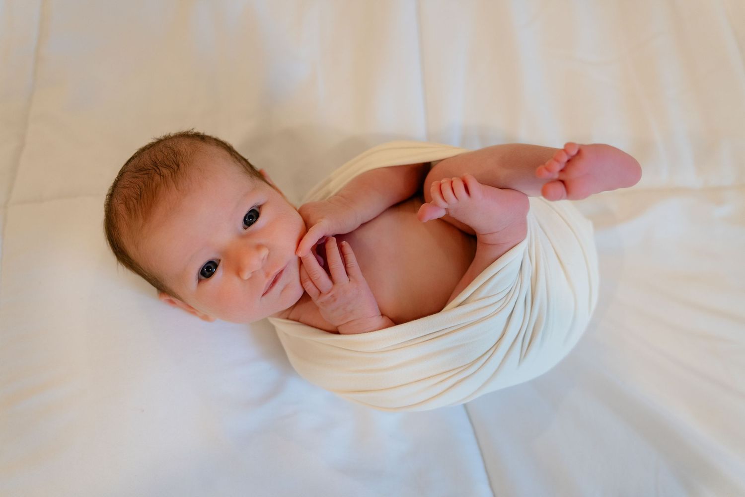 Newborn baby wrapped in a white swaddle blanket lying on a white surface, looking up with bright eyes.