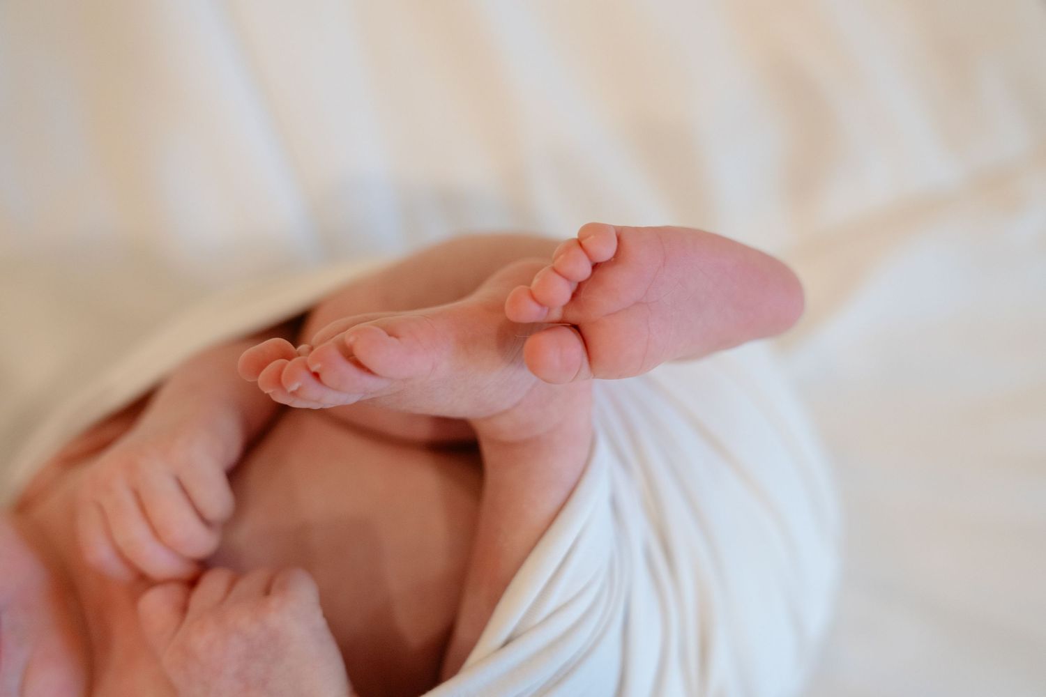Close-up of newborn baby feet and tiny hands wrapped in a soft white blanket on a white bed.
