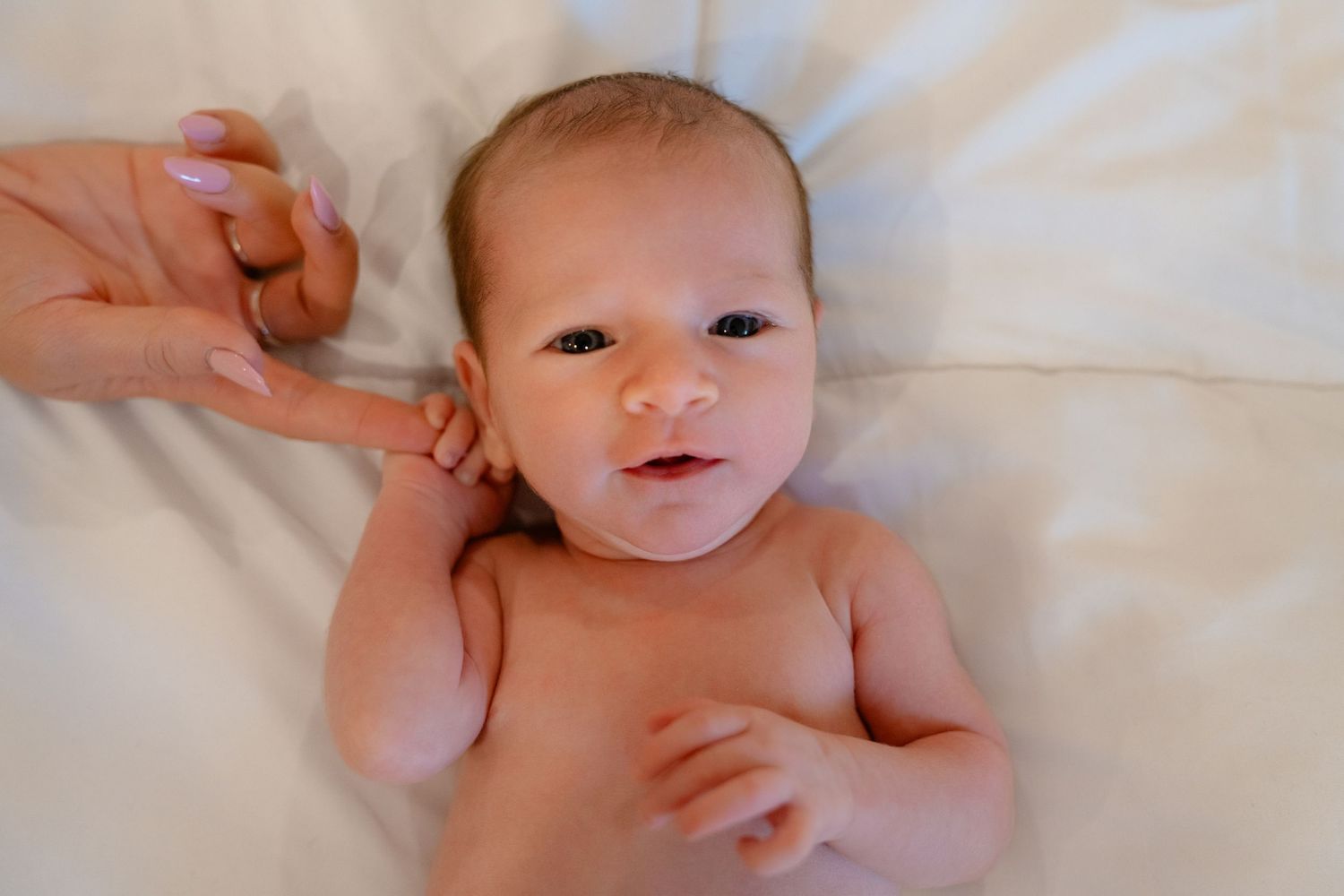 Newborn baby lying on white blanket, holding an adult finger, with alert eyes and a calm expression.