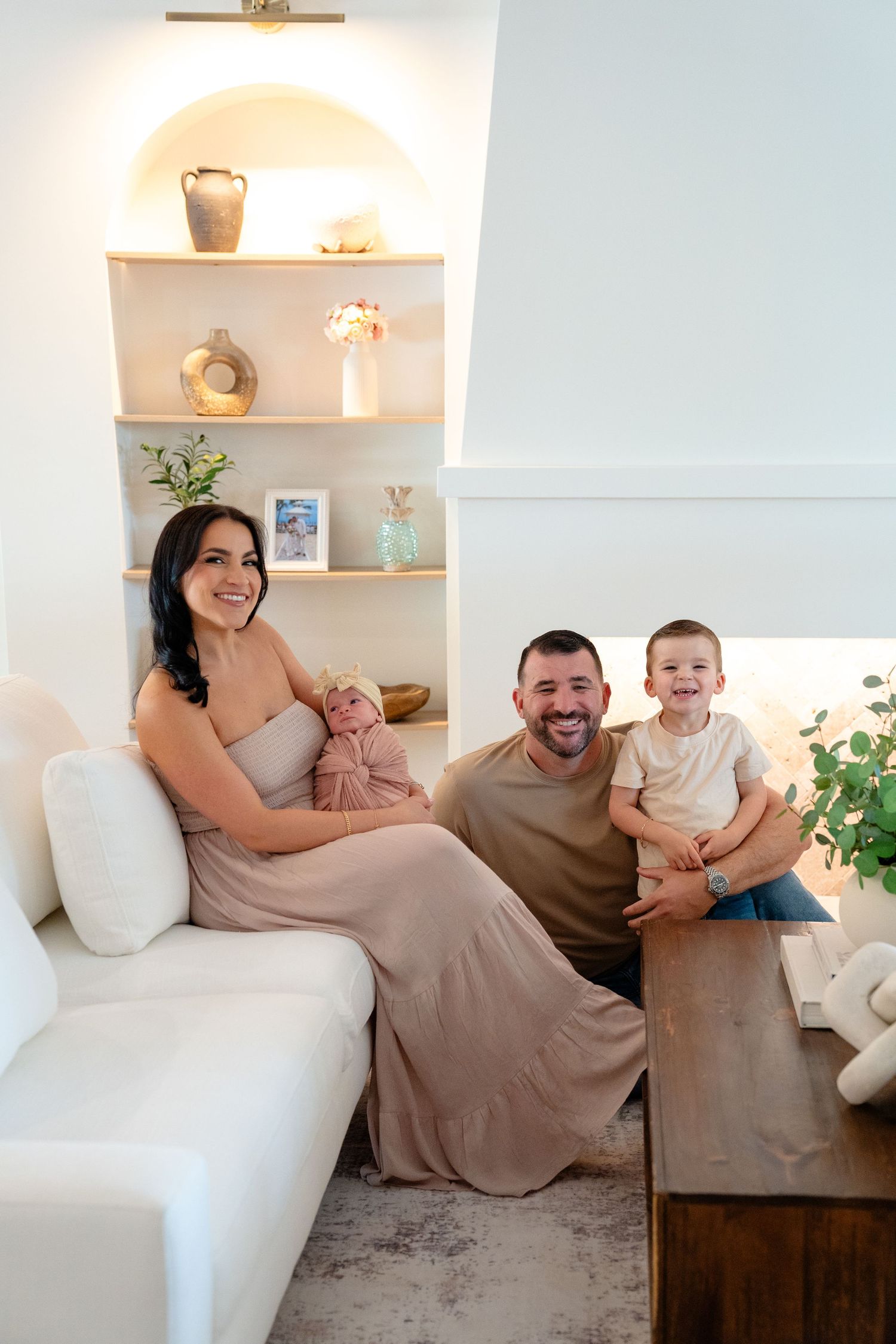 Happy family of four poses in a modern white living room, parents smiling with newborn baby and toddler son.