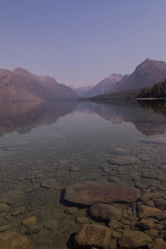 Crystal-clear mountain lake with rocky shoreline reflecting purple-hued peaks at dusk in Glacier National Park.