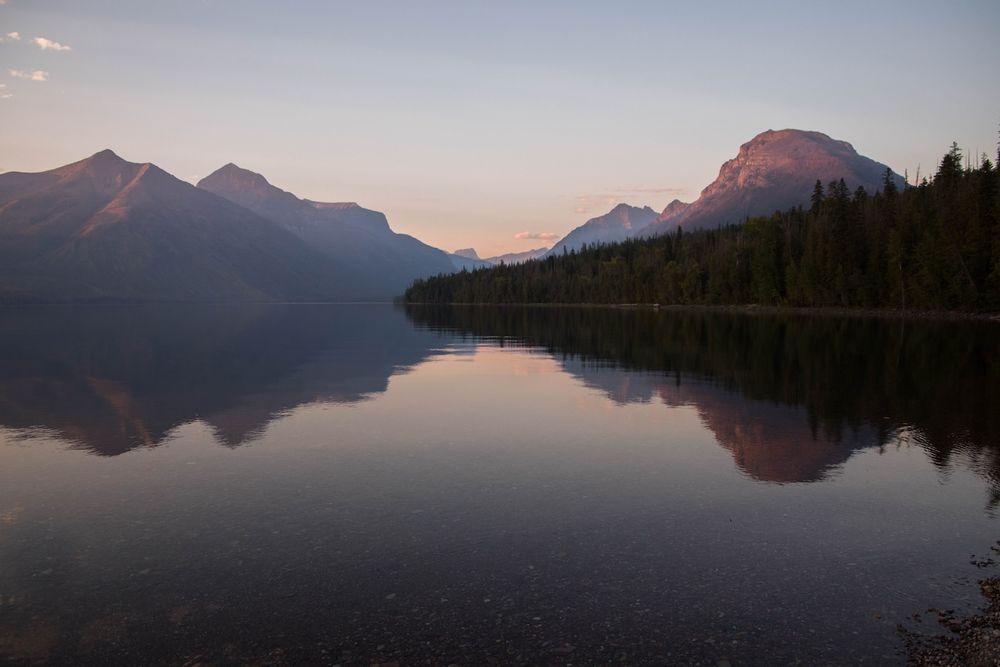 Serene mountain lake at sunset with pink-hued peaks reflecting on calm water, surrounded by dense evergreen forest.