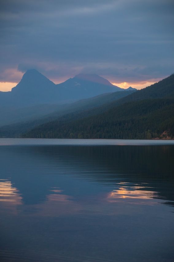 Serene mountain lake at dusk with misty peaks reflecting golden sunset light on calm water.