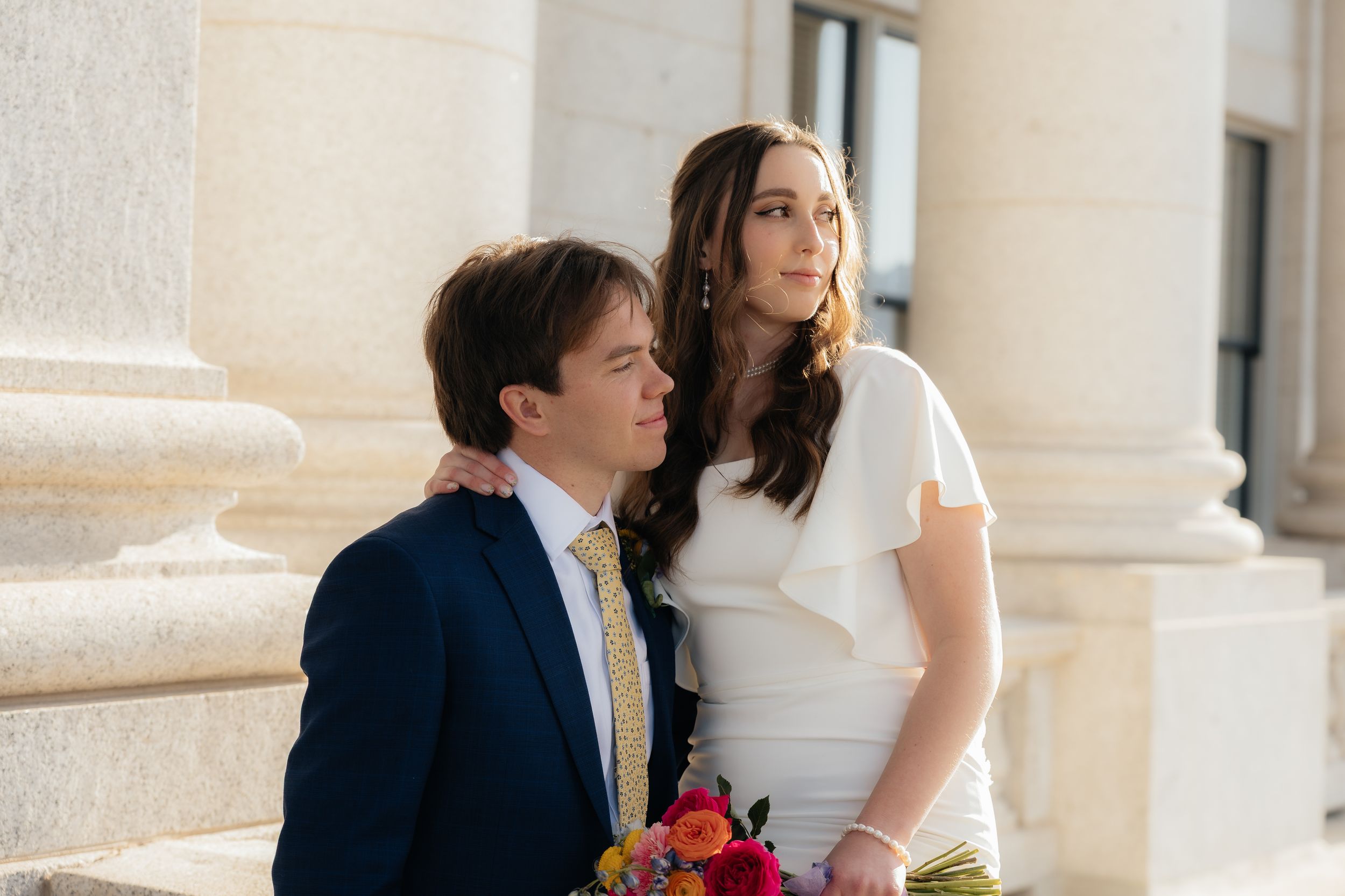 Bride in white dress holding colorful bouquet poses with groom in navy suit and yellow tie near stone columns.
