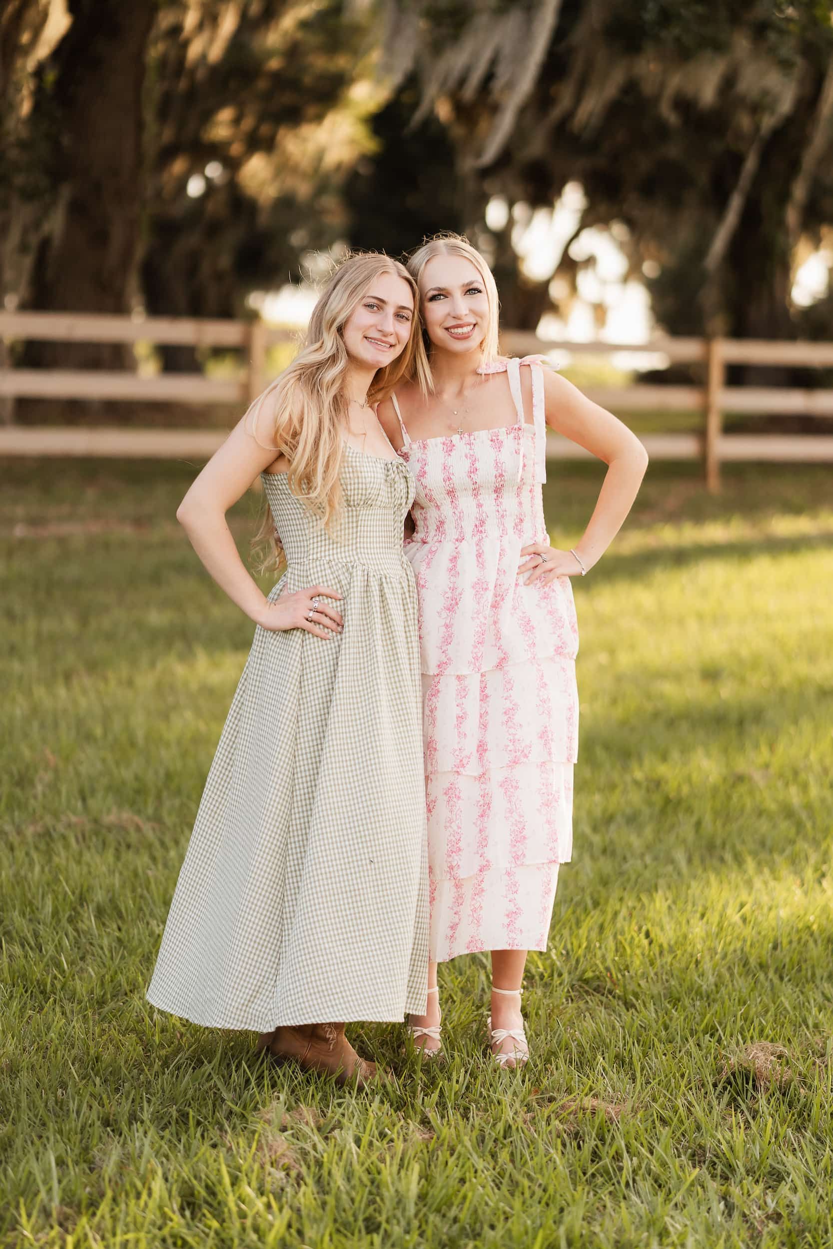 Two smiling women in floral summer dresses pose together on a green lawn near a wooden fence.