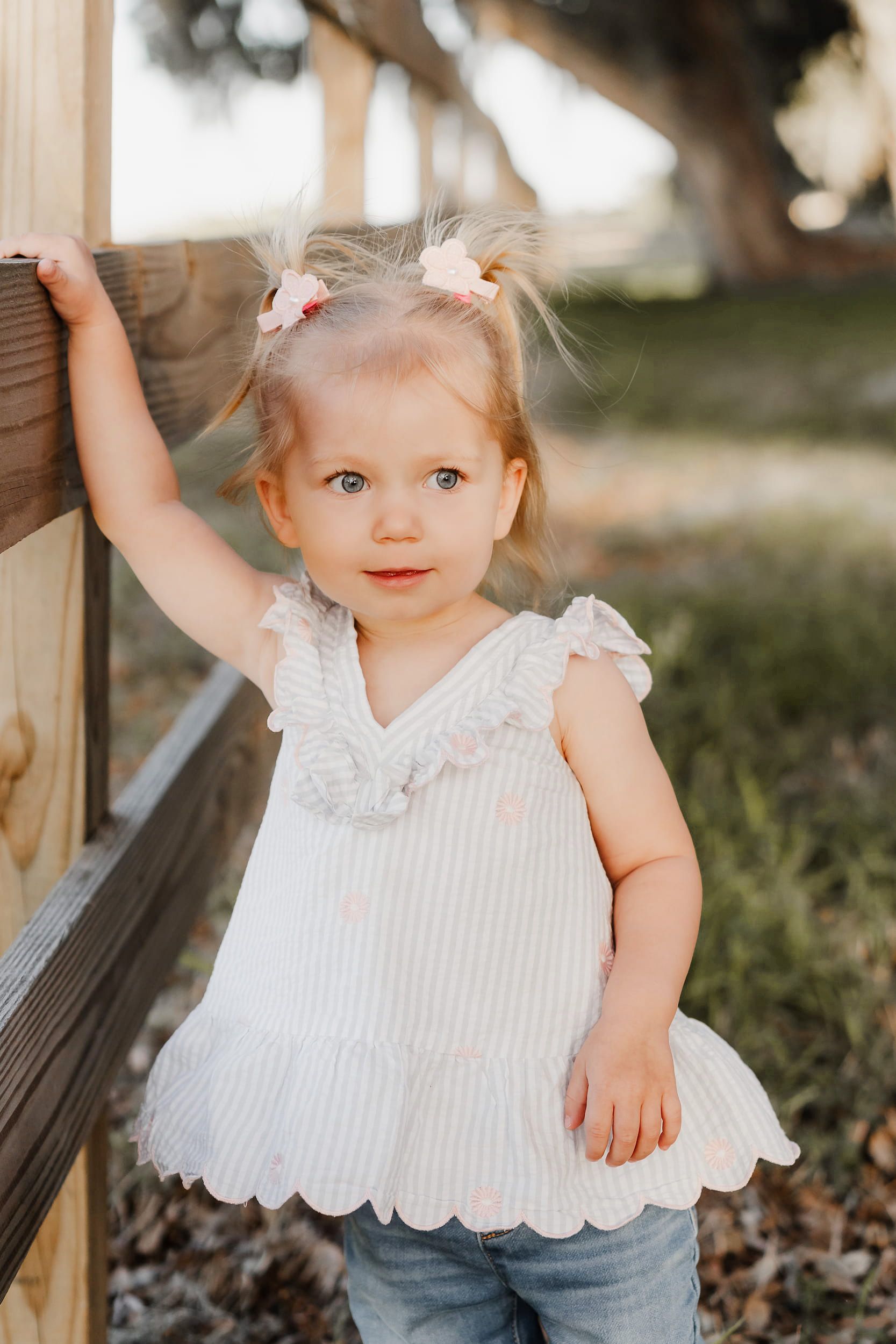Adorable toddler girl in white striped top with pink bows in pigtails, standing by a wooden fence outdoors.