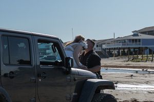 Couple sharing tender kiss seated on Jeep Wrangler bumper on sandy beach near coastal buildings.