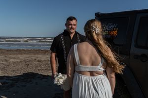 Groom in black shirt smiling at bride in backless white dress near decorated Jeep on beach.