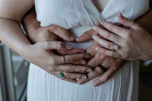 Close-up of couple's hands forming heart shape on bride's belly wearing wedding and turquoise rings.