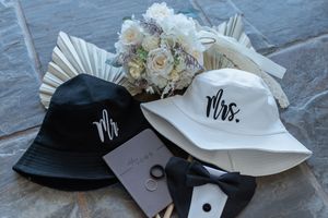 Mr and Mrs bucket hats with white floral bouquet, wedding rings, and bow tie flat lay display.